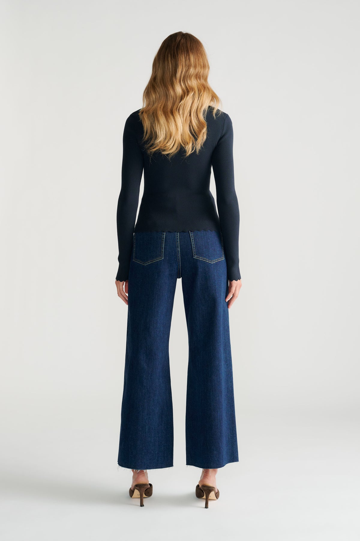 Back view of woman with loose wavy hair wearing a fitted navy scalloped top and wide-leg dark denim jeans with heels.