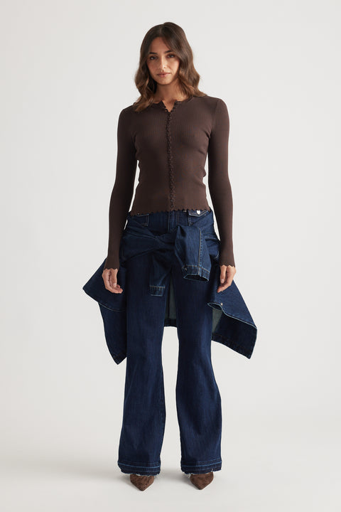 Woman wearing a fitted chocolate brown scalloped button long sleeve top with dark wide-leg denim jeans and brown pointed shoes