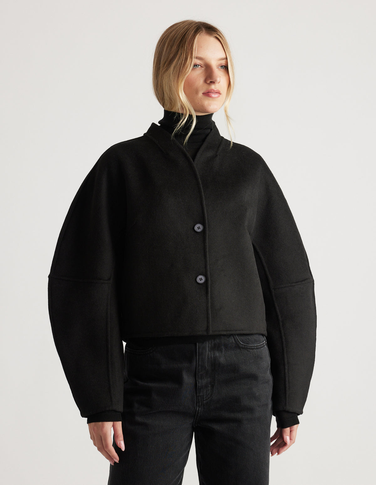 Woman wearing a black cropped jacket with voluminous barrel sleeves and a sleek high collar, paired with black pants