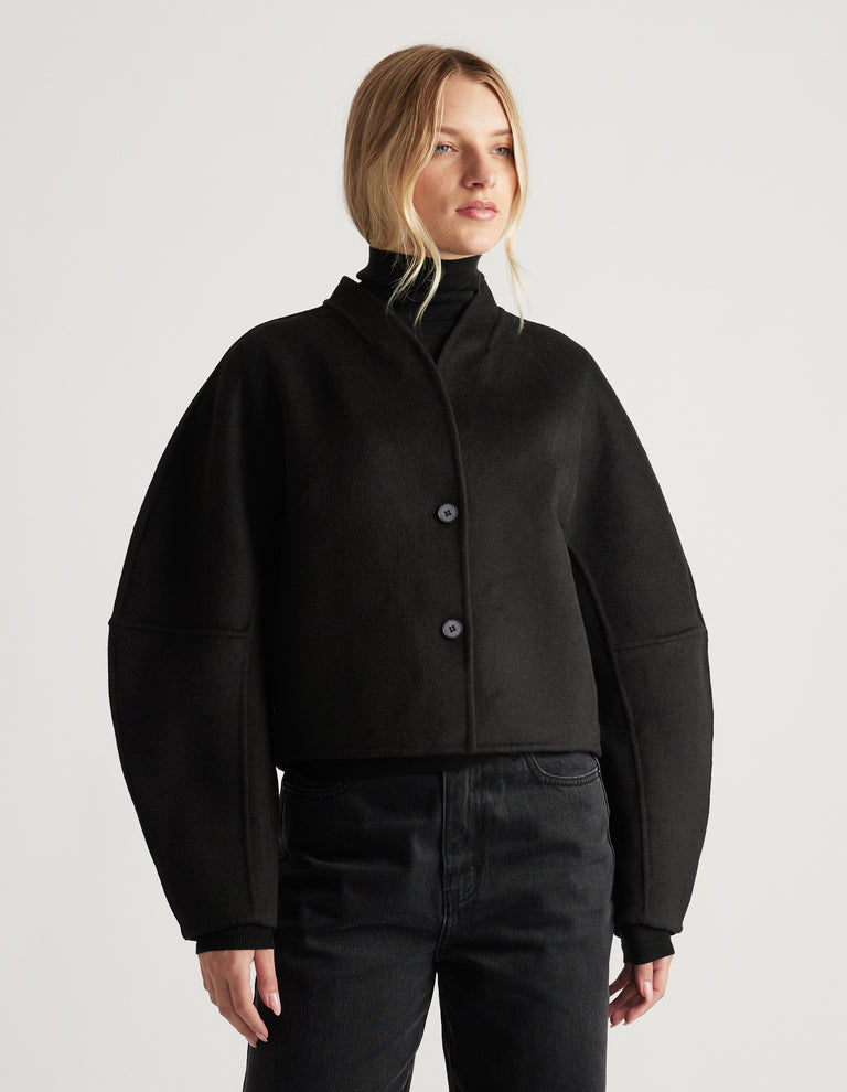 Woman wearing a black cropped jacket with voluminous barrel sleeves and a sleek high collar, paired with black pants
