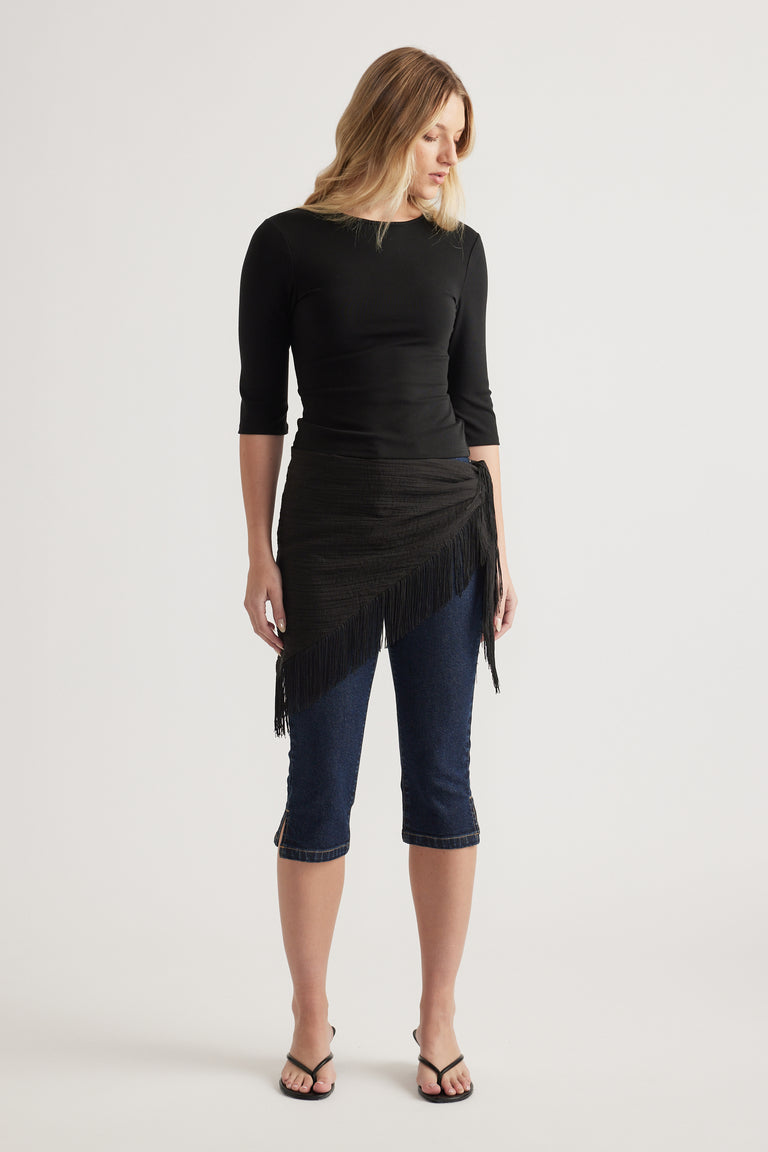 Woman wearing a fitted black three-quarter sleeve top with a fringed wrap skirt over cropped jeans