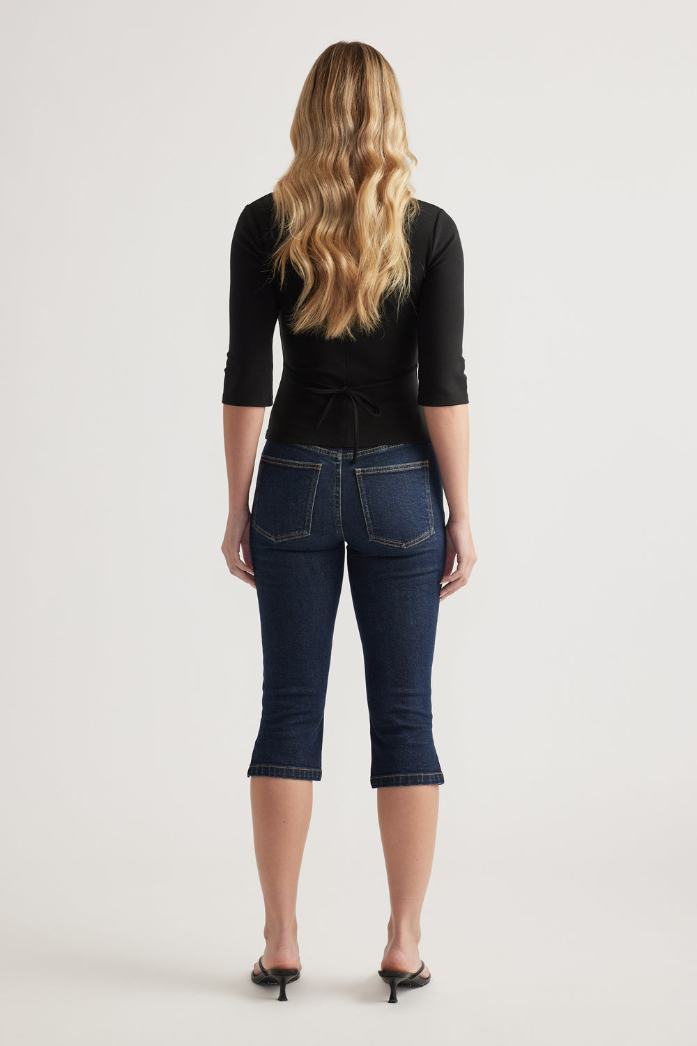 Woman wearing a fitted black three quarter sleeve top with back waist tie and dark cropped jeans