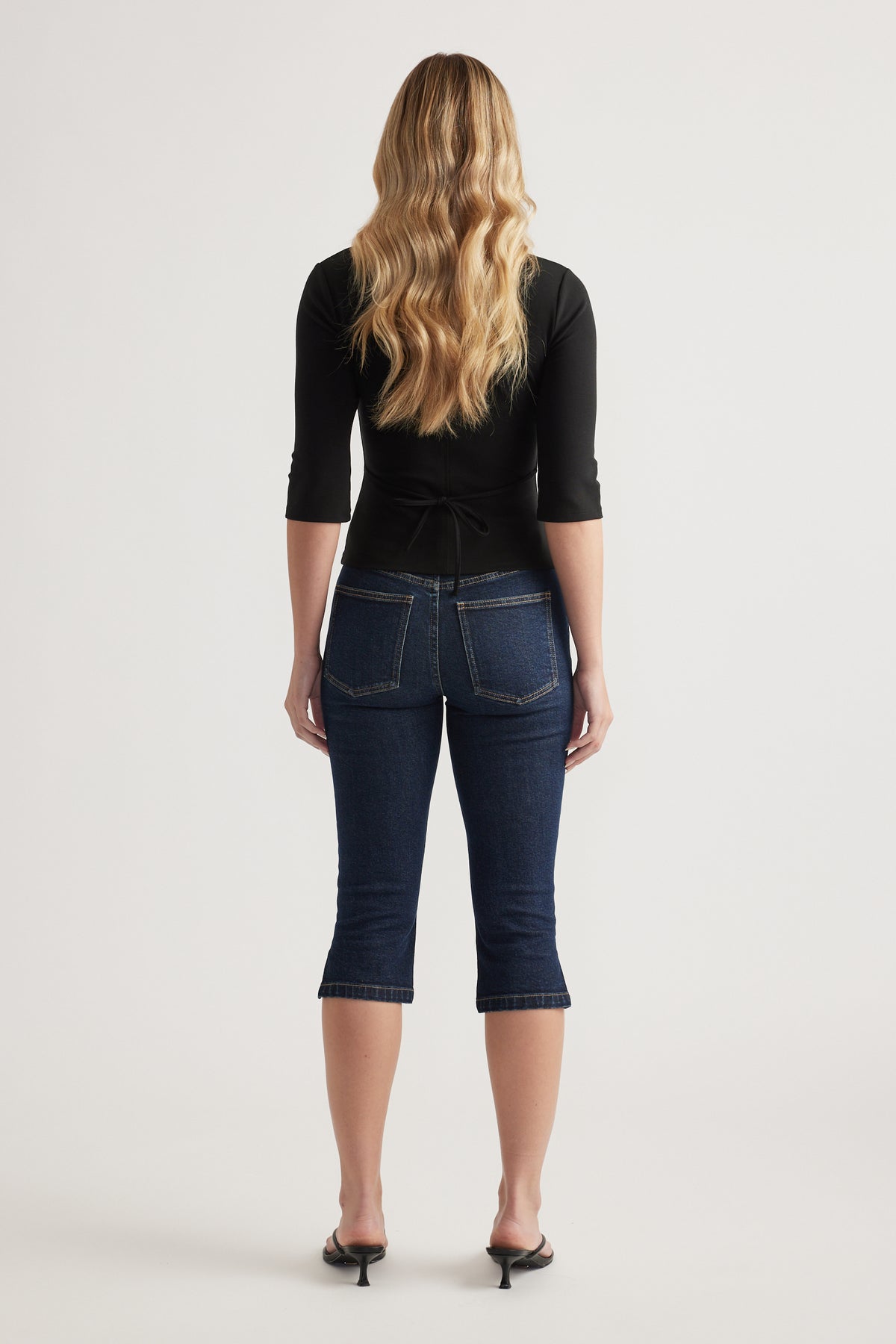 Woman wearing a fitted black three quarter sleeve top with back waist tie and dark cropped jeans