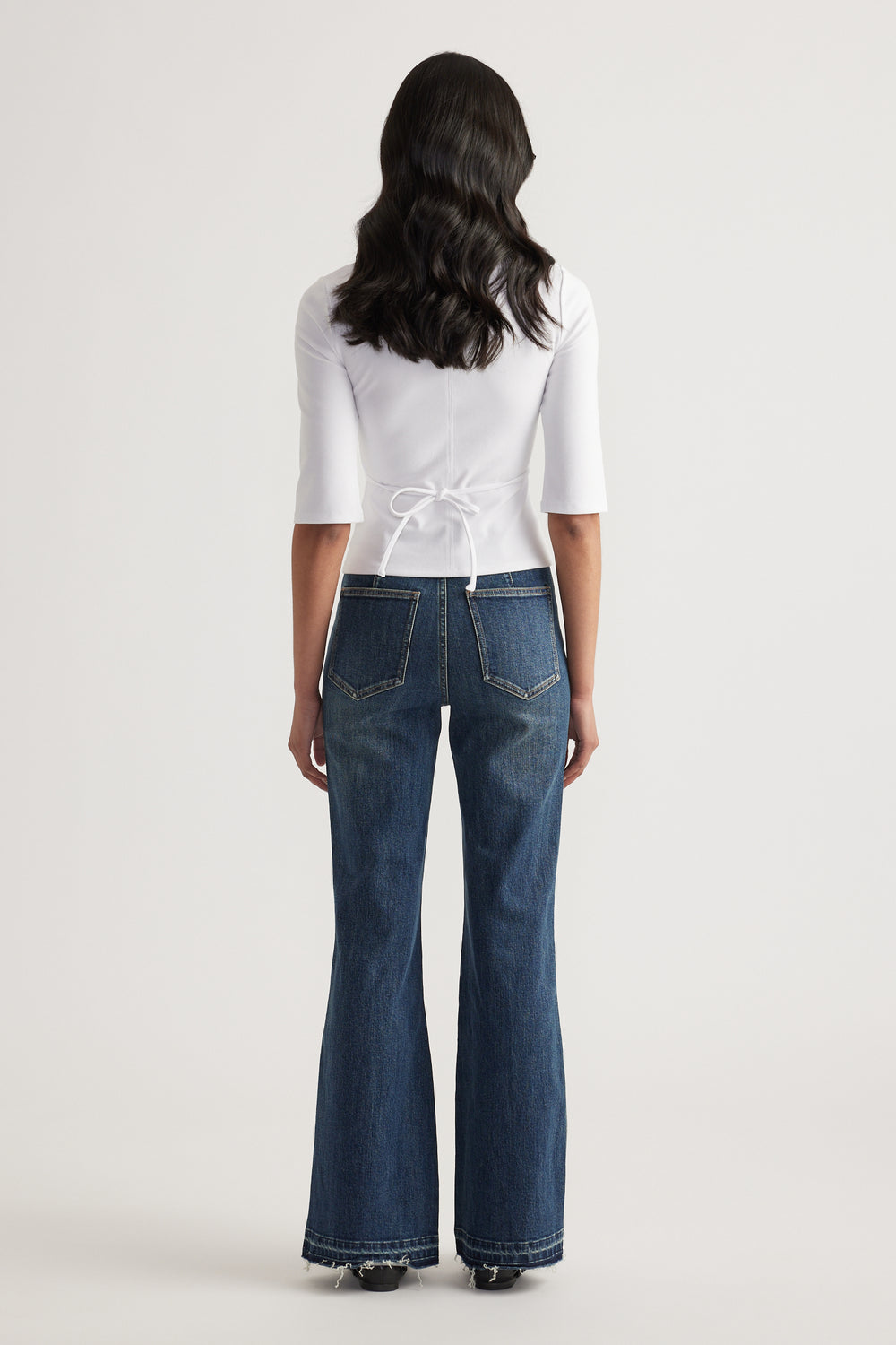 Back view of a woman wearing a fitted white three-quarter sleeve top with waist tie and wide-leg blue jeans