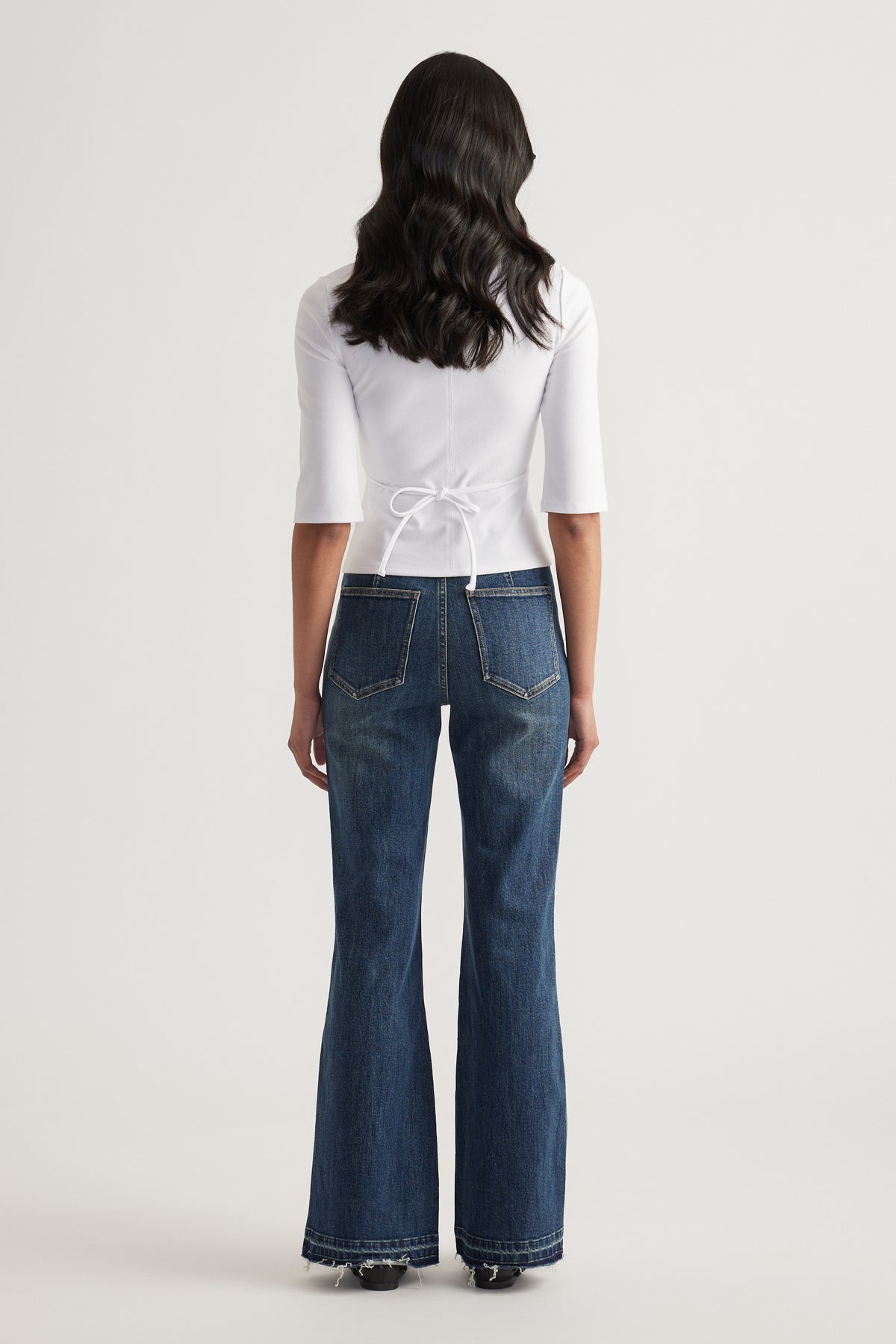 Back view of a woman wearing a fitted white three-quarter sleeve top with waist tie and wide-leg blue jeans
