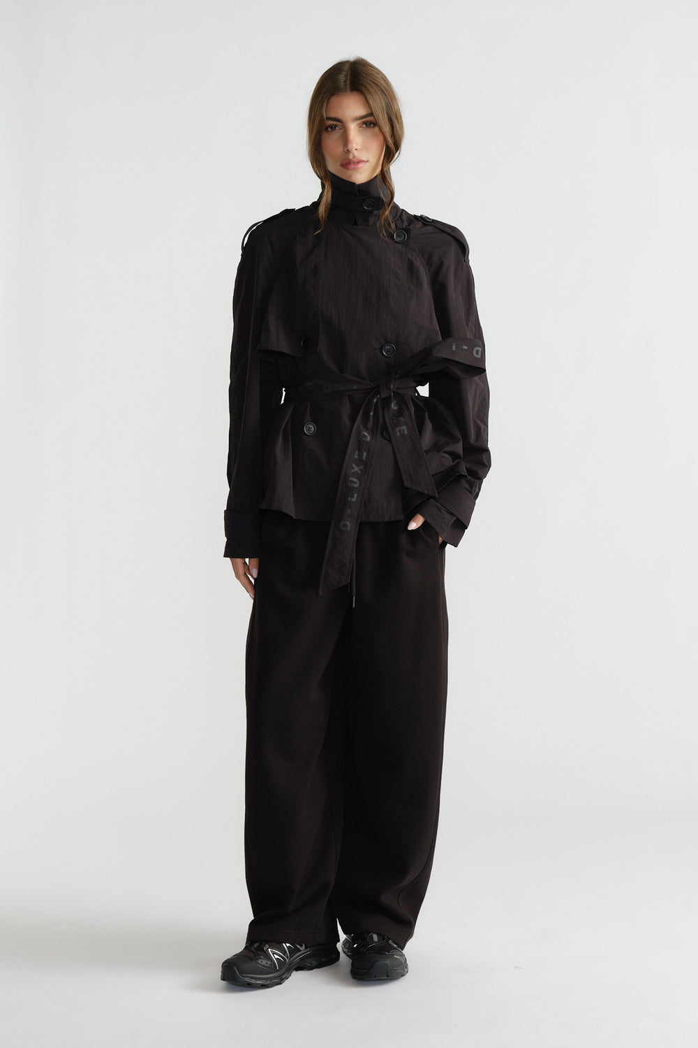 Woman wearing a black belted jacket with structured shoulders paired with loose black barrel track pants and black sneakers