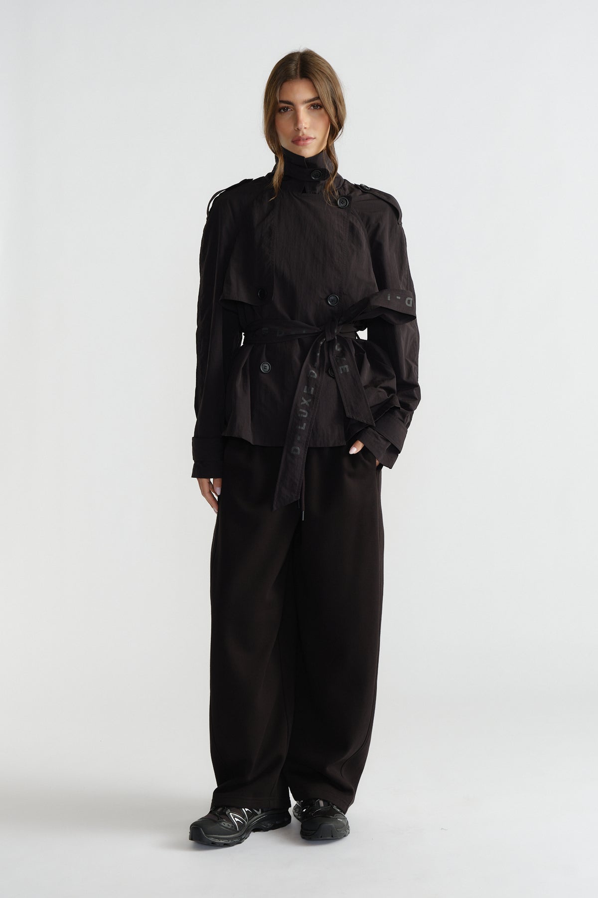 Woman wearing a black belted jacket with structured shoulders paired with loose black barrel track pants and black sneakers