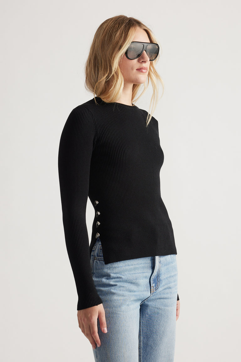 Woman wearing a fitted black ribbed long sleeve top with metal side detailing paired with light wash blue jeans and sunglasses
