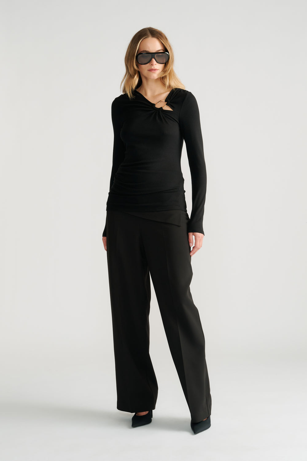 Woman wearing a black long-sleeve top with metal detail paired with wide-leg black pants and black heels.