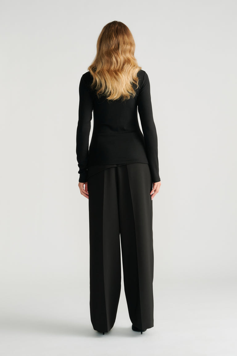 Woman wearing a slim-fit black top with metal accents paired with wide-leg tailored black pants, shown from behind.