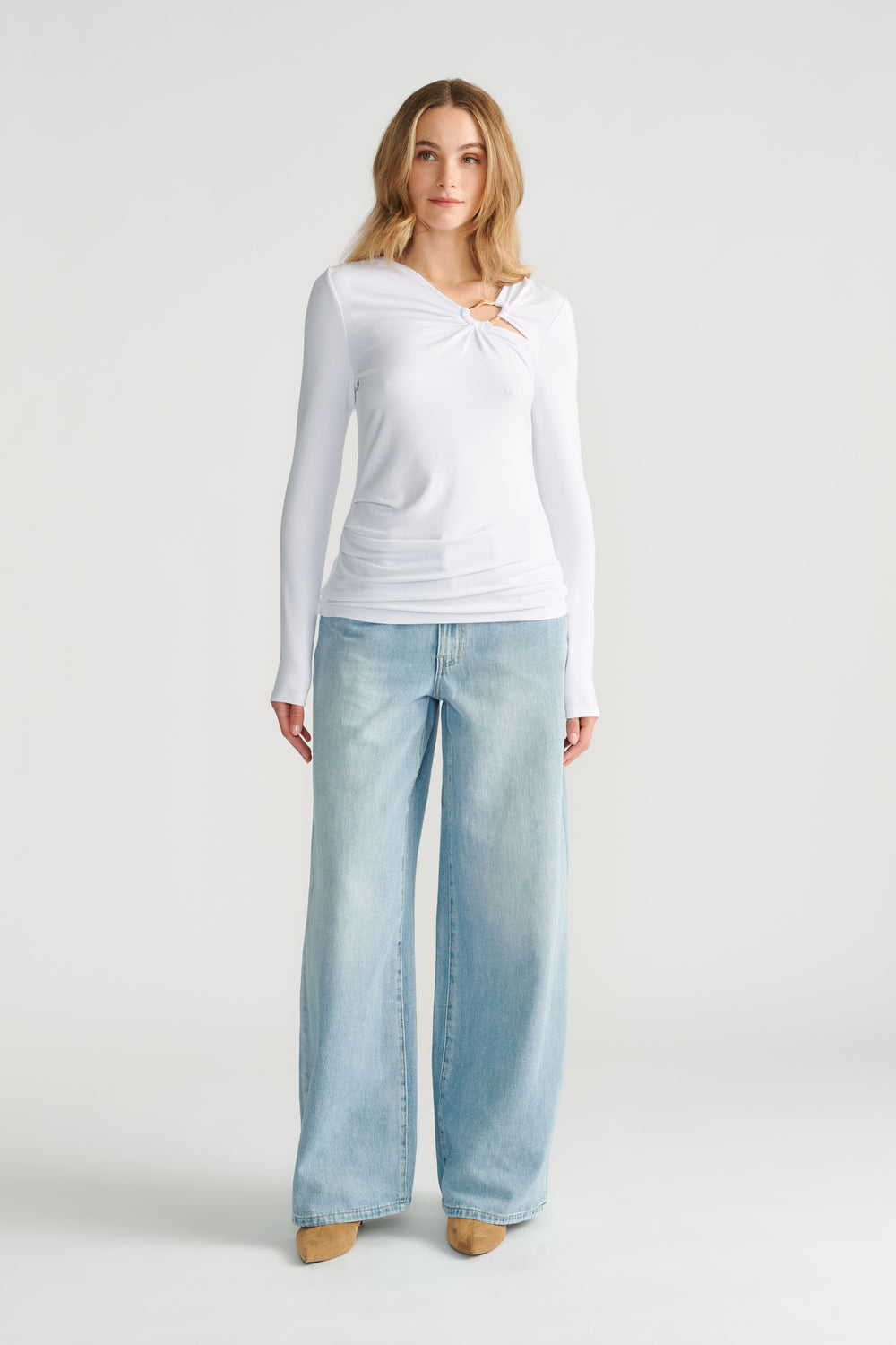 Woman wearing a white long-sleeve top with a metal ring detail paired with wide-leg light wash denim jeans and tan shoes.