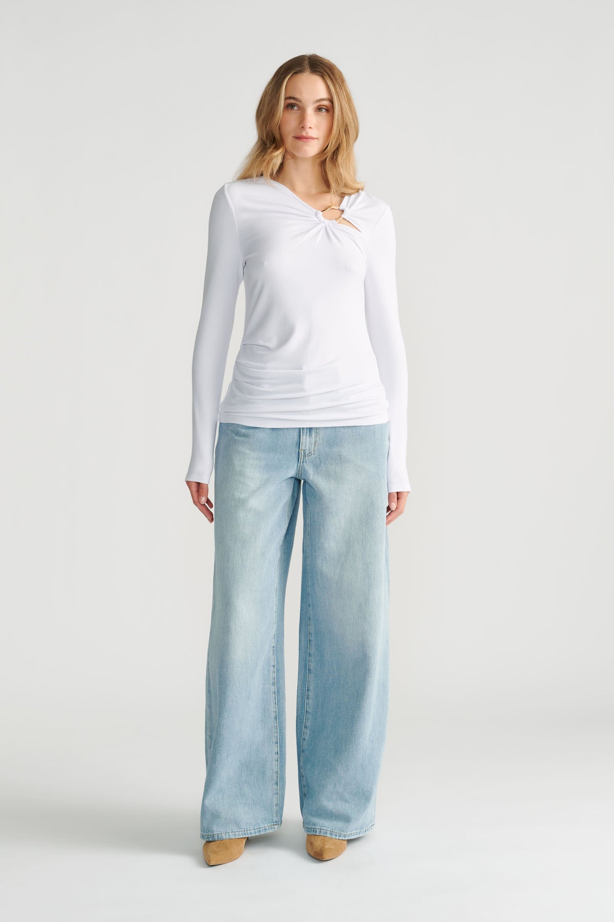 Woman wearing a white long-sleeve top with a metal ring detail paired with wide-leg light wash denim jeans and tan shoes.