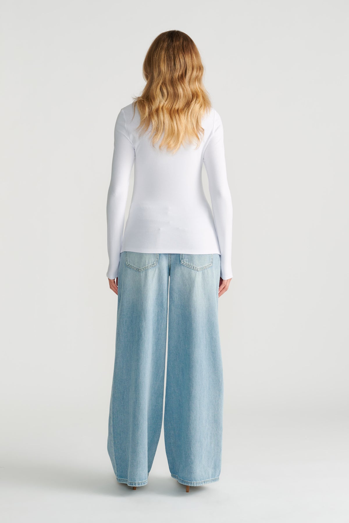 Woman wearing a fitted white long-sleeve top paired with light wash wide-leg denim jeans, shown from behind.