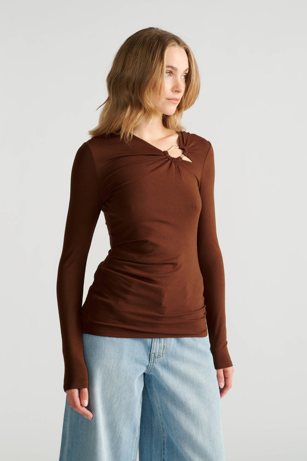 Woman wearing a fitted long-sleeve brown top with metal detail at neckline and light blue wide-leg jeans