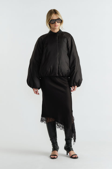 Woman wearing an oversized black bomber jacket with balloon sleeves over a black lace-trim midi skirt and heeled boots