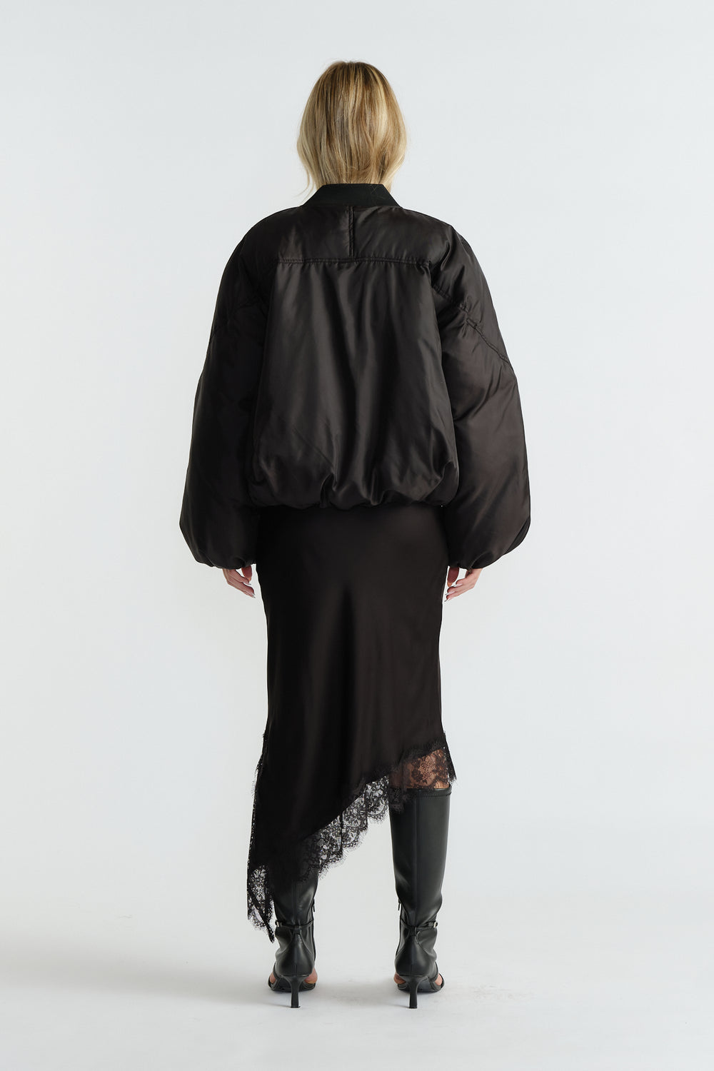 Back view of woman wearing a black oversized bomber jacket paired with an asymmetric lace-hem skirt and black heeled boots
