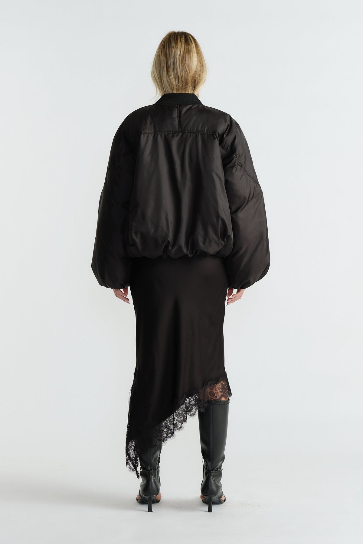 Back view of woman wearing a black oversized bomber jacket paired with an asymmetric lace-hem skirt and black heeled boots