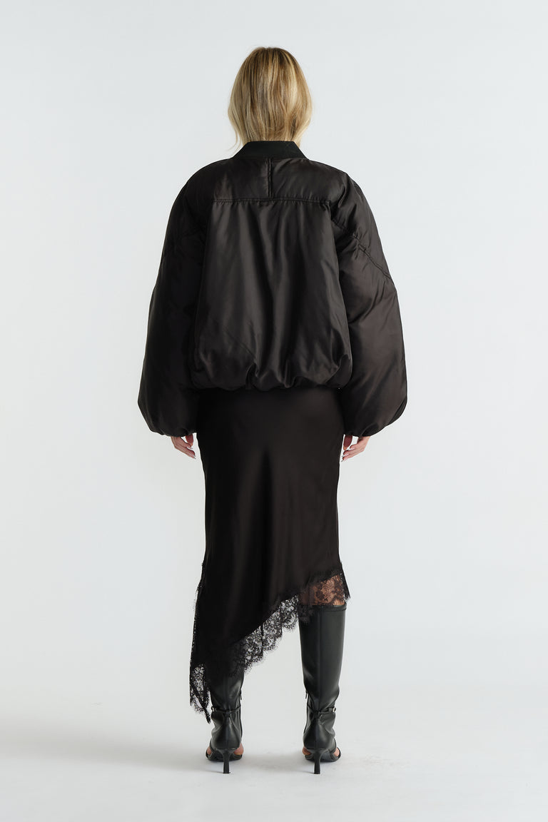 Back view of woman wearing a black oversized bomber jacket paired with an asymmetric lace-hem skirt and black heeled boots