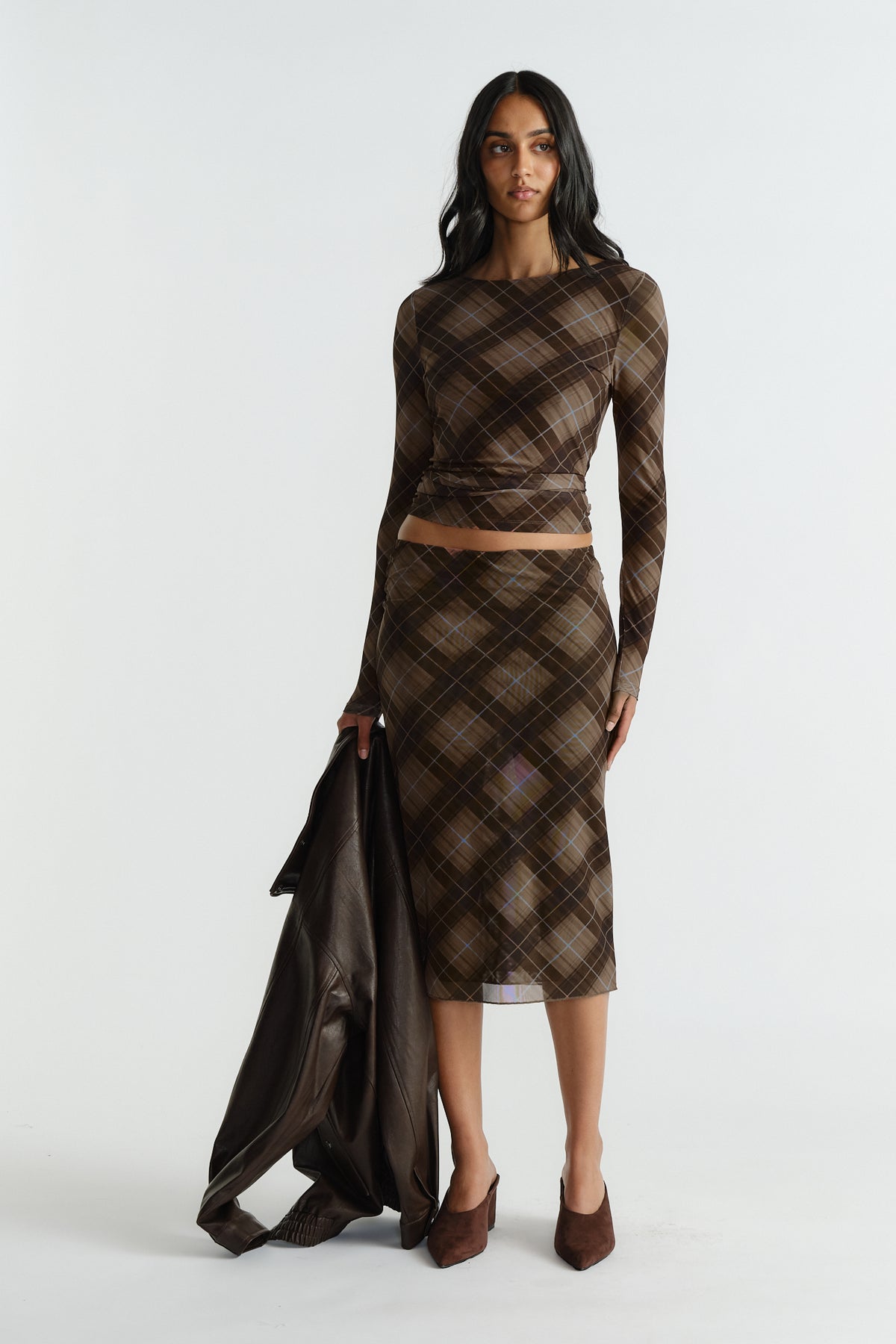 Woman wearing a fitted brown plaid mesh top and matching midi skirt, holding a dark leather jacket and wearing brown heels.
