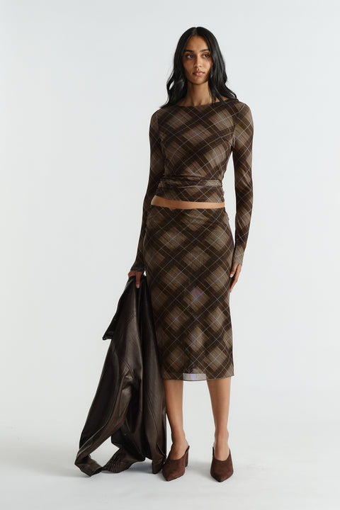 Woman wearing a fitted brown plaid mesh top and matching midi skirt, holding a dark leather jacket and wearing brown heels.