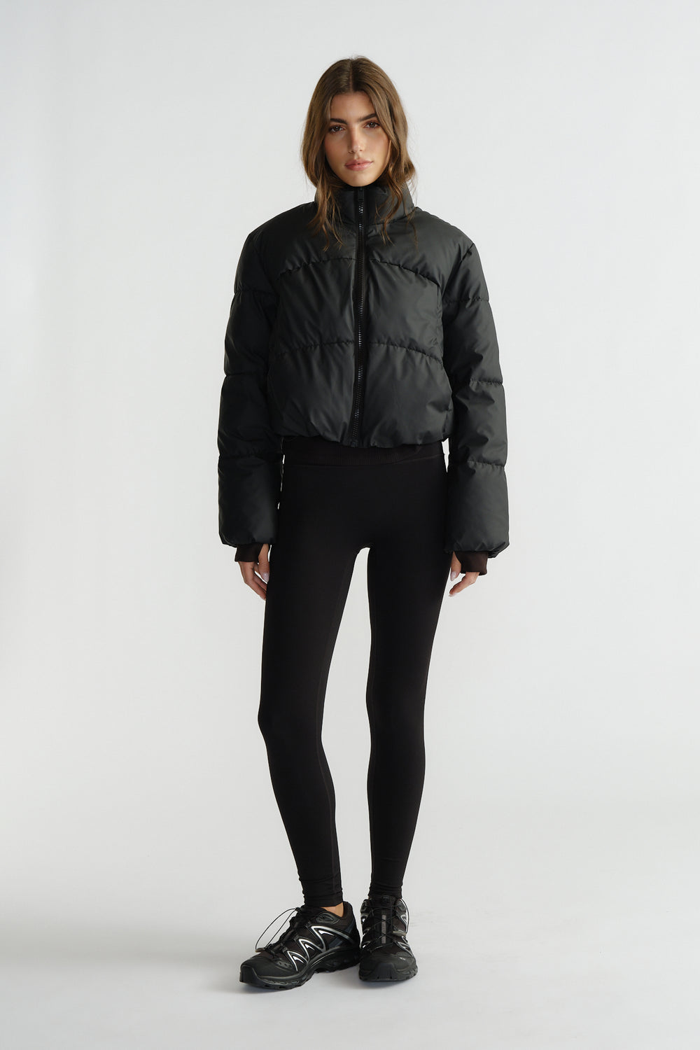 Woman wearing a black cropped puffer jacket with a high collar, paired with fitted black leggings and sneakers