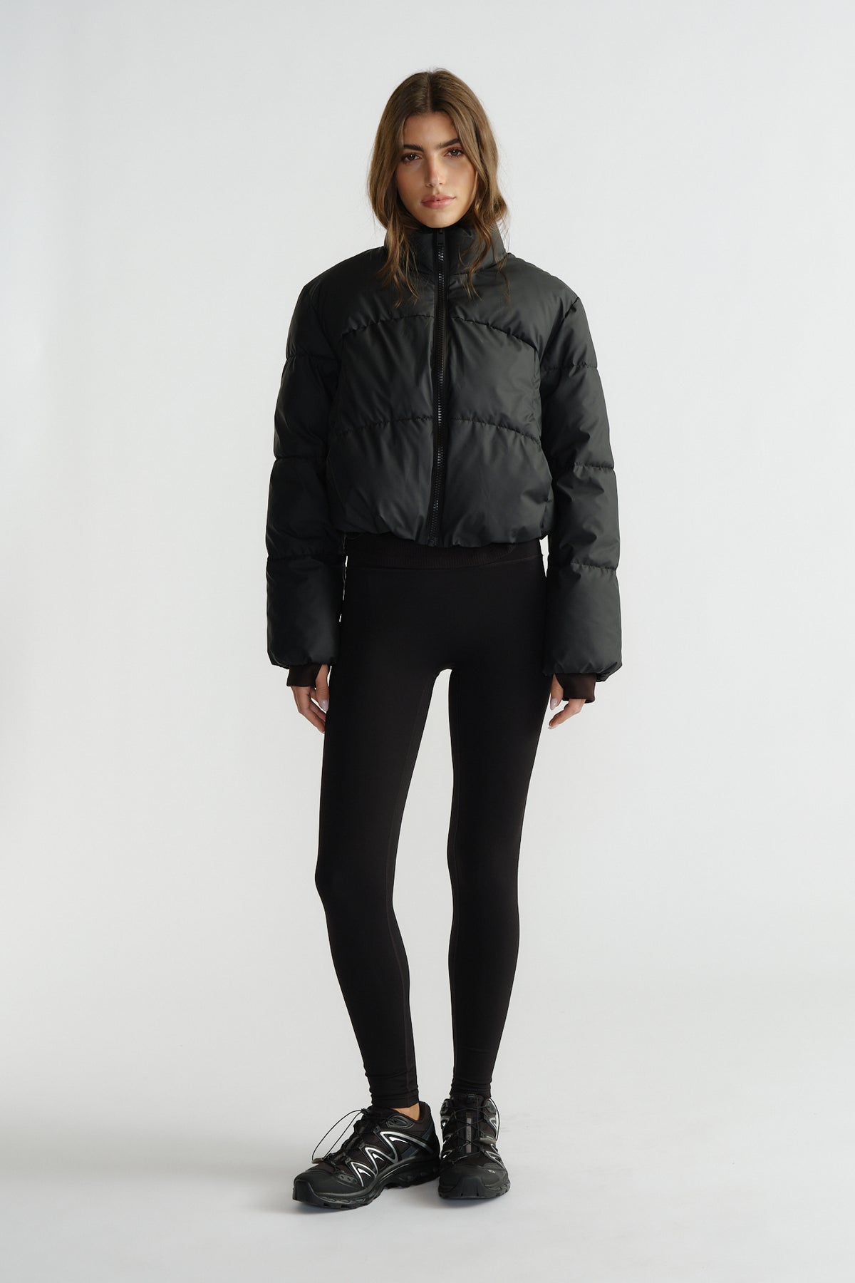 Woman wearing a black cropped puffer jacket with a high collar, paired with fitted black leggings and sneakers