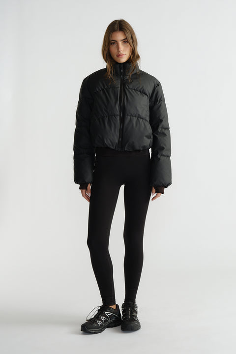 Woman wearing a black cropped puffer jacket with a high collar, paired with fitted black leggings and sneakers