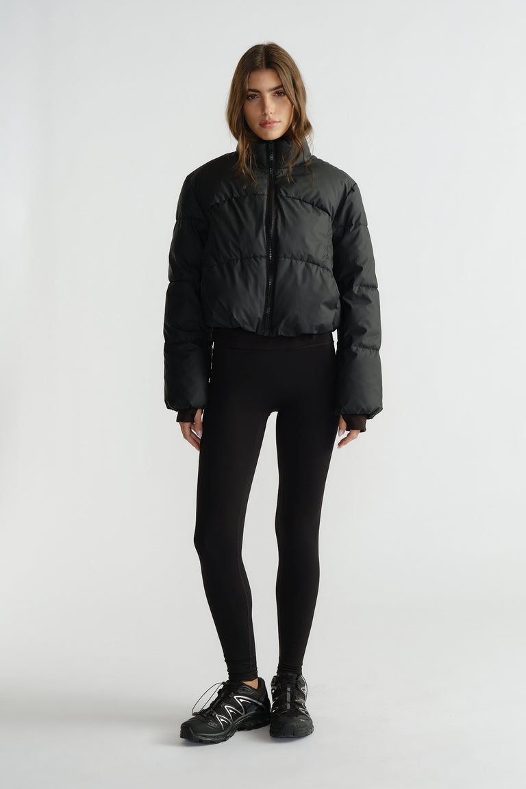 Woman wearing a black cropped puffer jacket with a high collar, paired with fitted black leggings and sneakers
