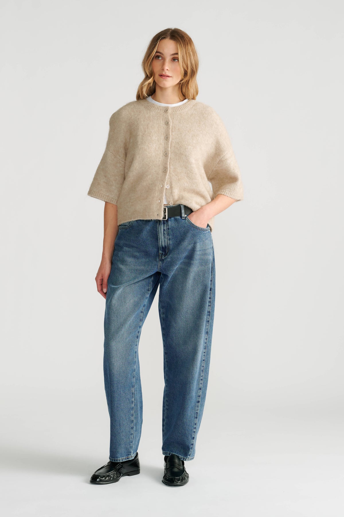 Woman wearing a soft beige boxy knit cardigan with button details paired with relaxed fit blue jeans and black shoes.