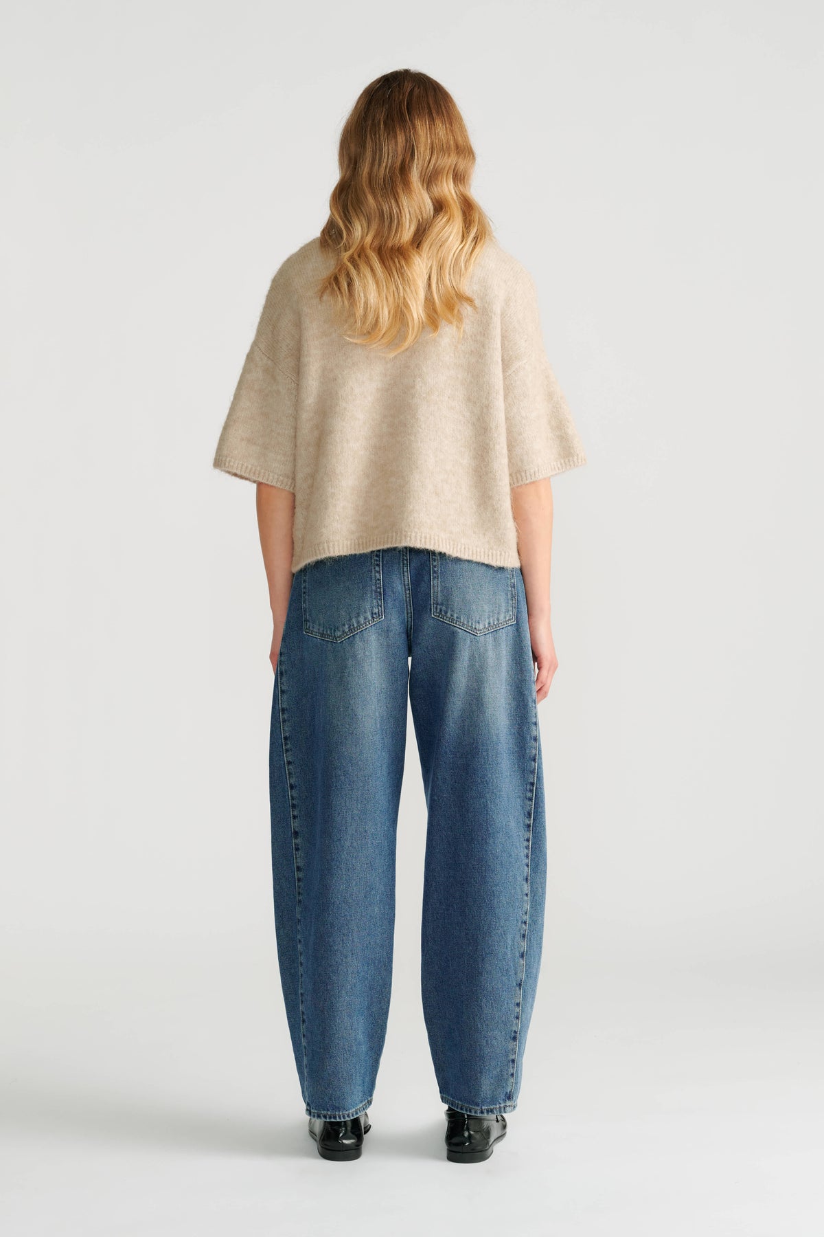 Woman wearing a soft beige boxy knit cardigan paired with relaxed-fit blue denim jeans and black shoes, seen from behind.