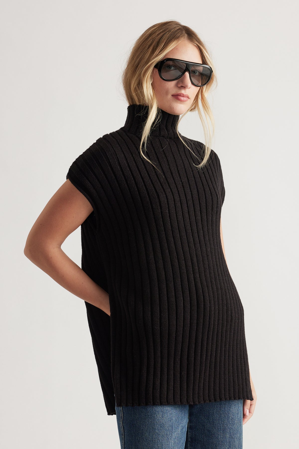 Woman wearing a black ribbed knit sleeveless tunic with a high neck paired with blue jeans and oversized sunglasses