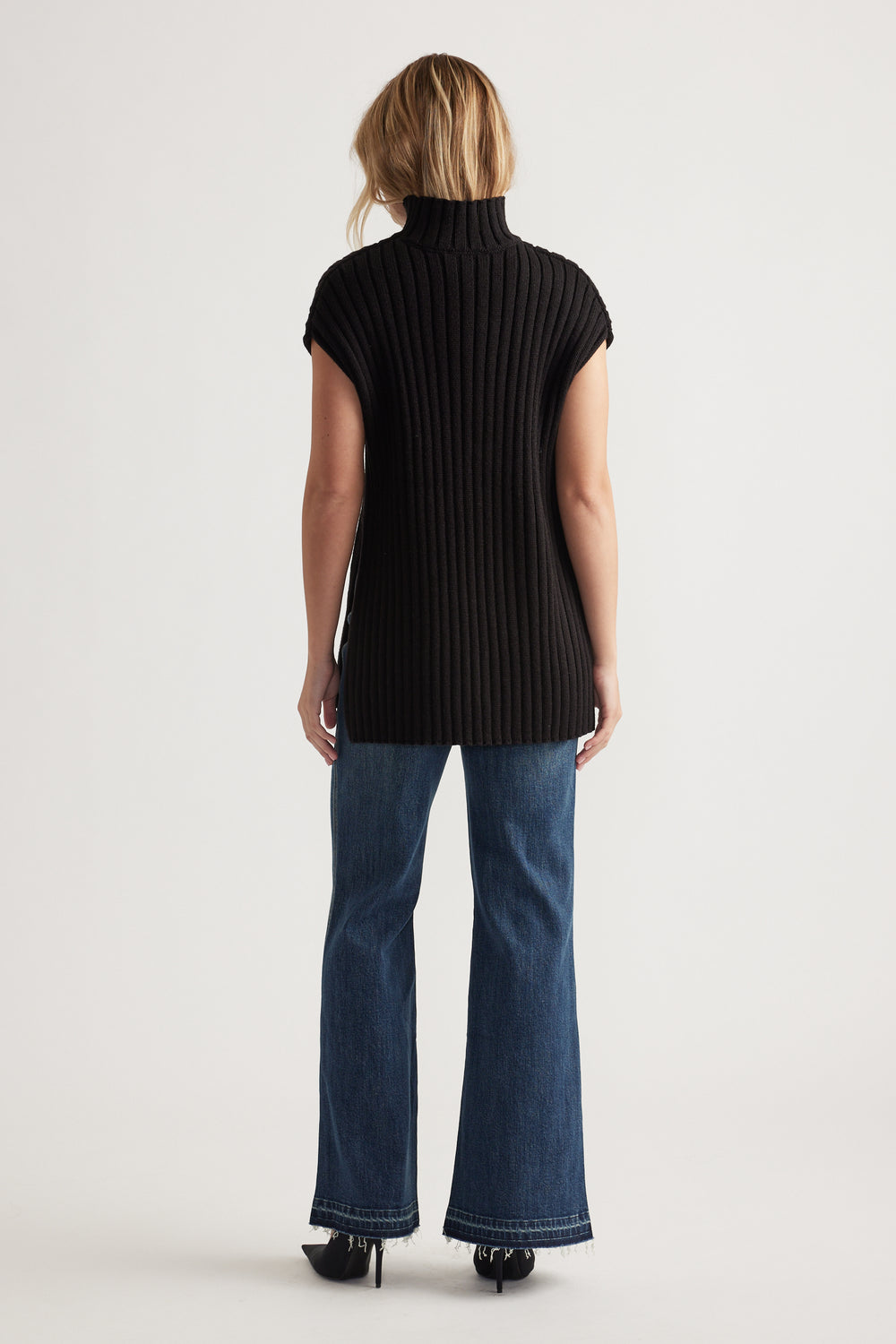 Woman wearing a long black ribbed knit tunic with high neck and cap sleeves paired with dark flared jeans and heels