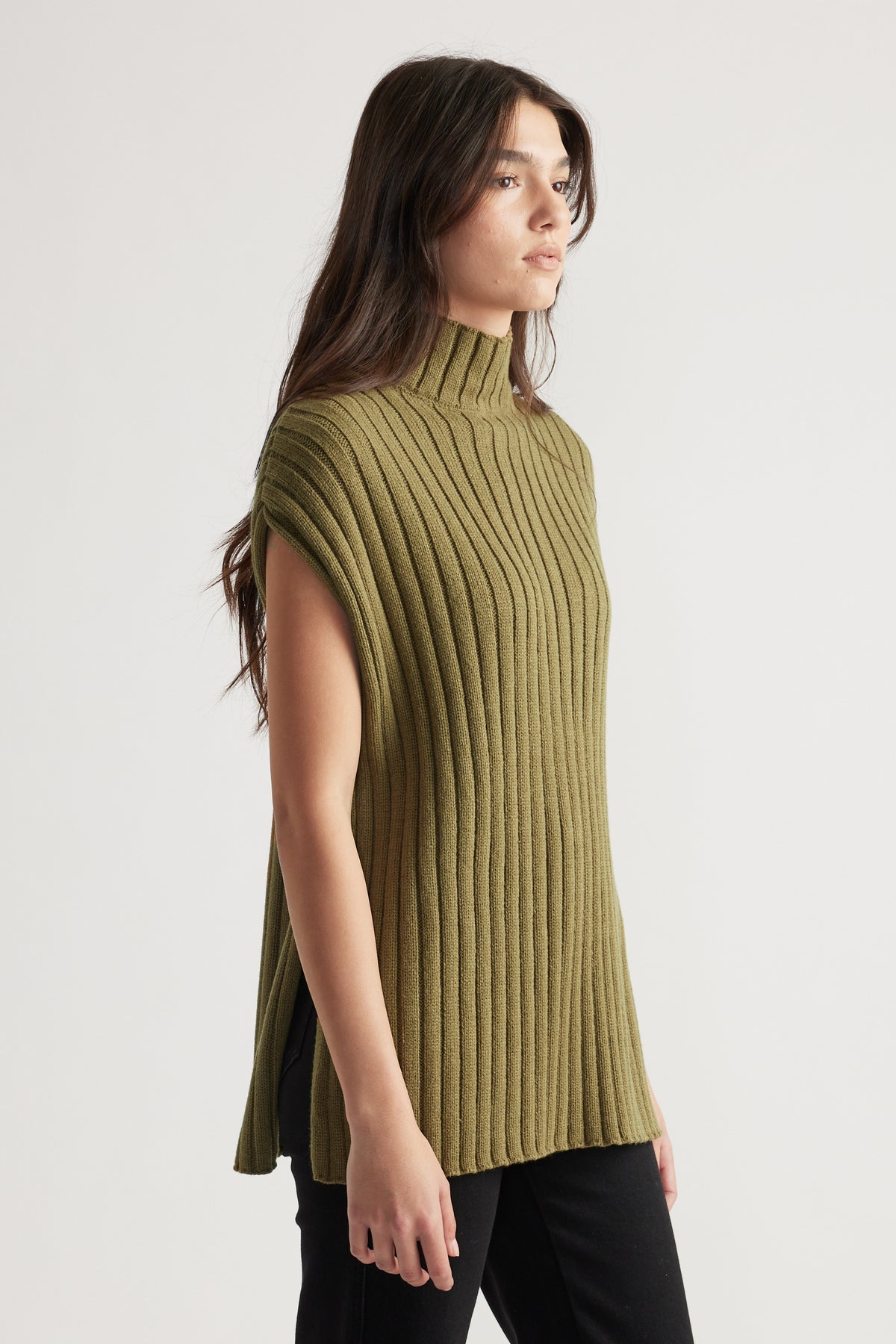 Woman wearing a sleeveless ribbed knit tunic with a high neck in olive green paired with black pants