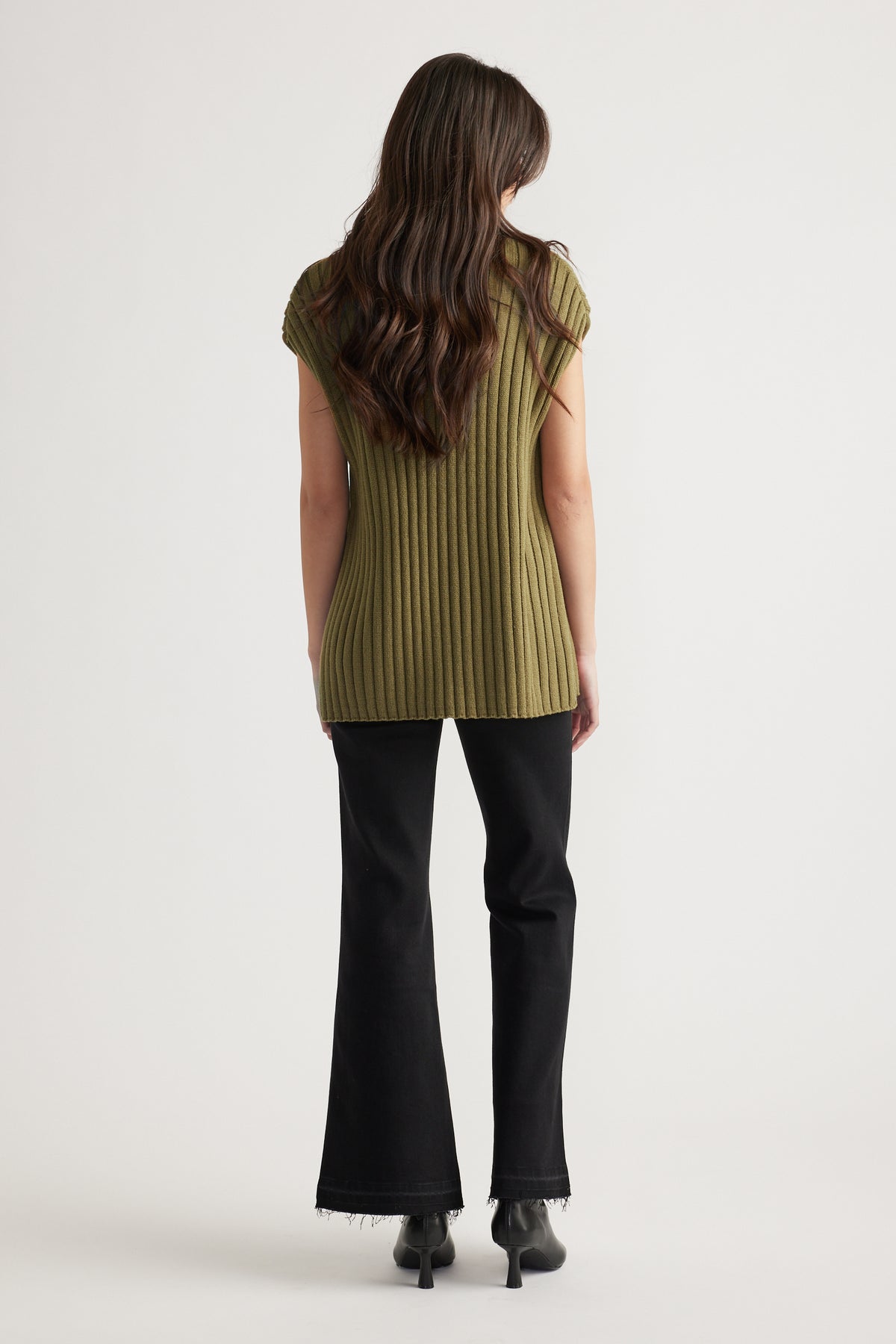 Woman with long wavy hair wearing an olive ribbed knit tunic paired with black flared jeans and heeled boots, back view.