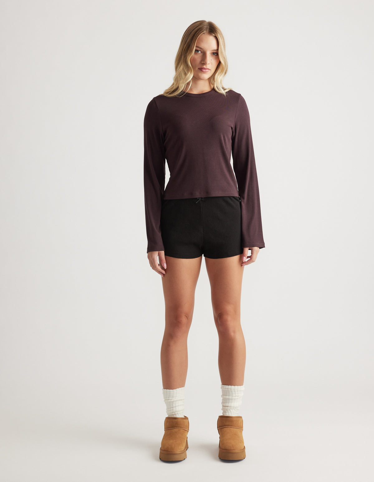 Woman wearing a soft ribbed long sleeve top in deep rhubarb paired with black shorts and cozy ankle boots