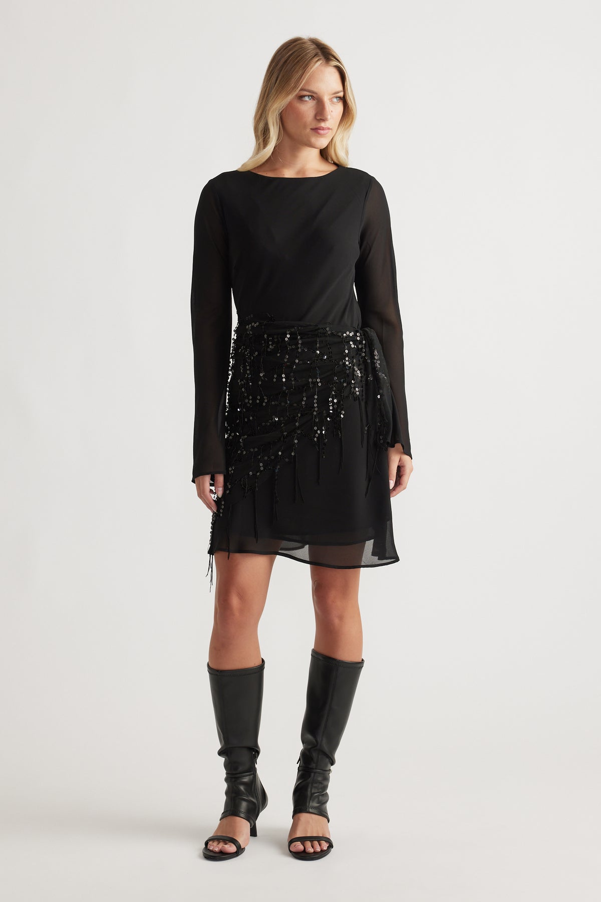 Woman wearing a black long-sleeve dress with sequin wrap detail and knee-high black open-toe boots.
