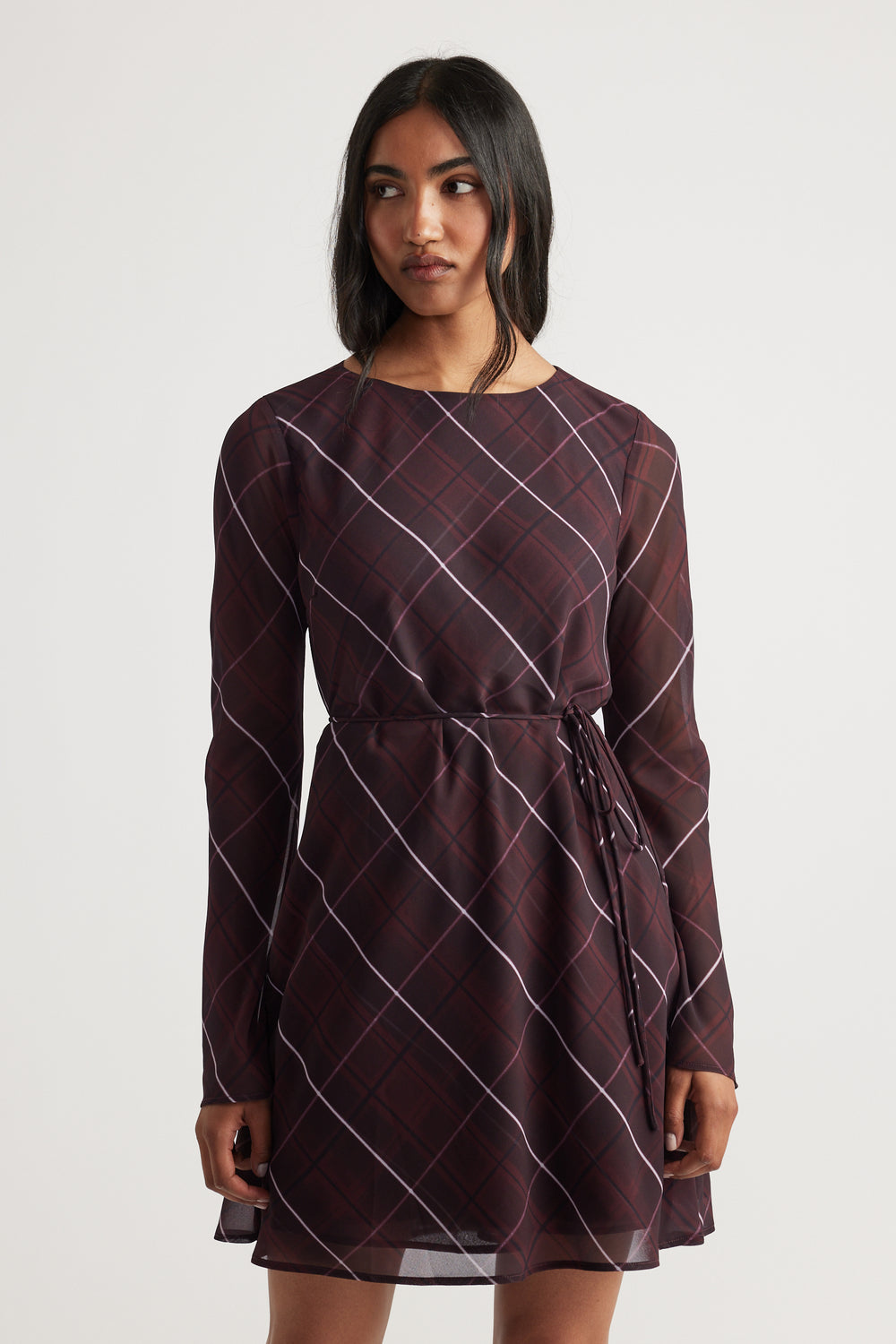 Woman wearing a long-sleeve dark plaid dress with a waist tie, standing against a plain light background