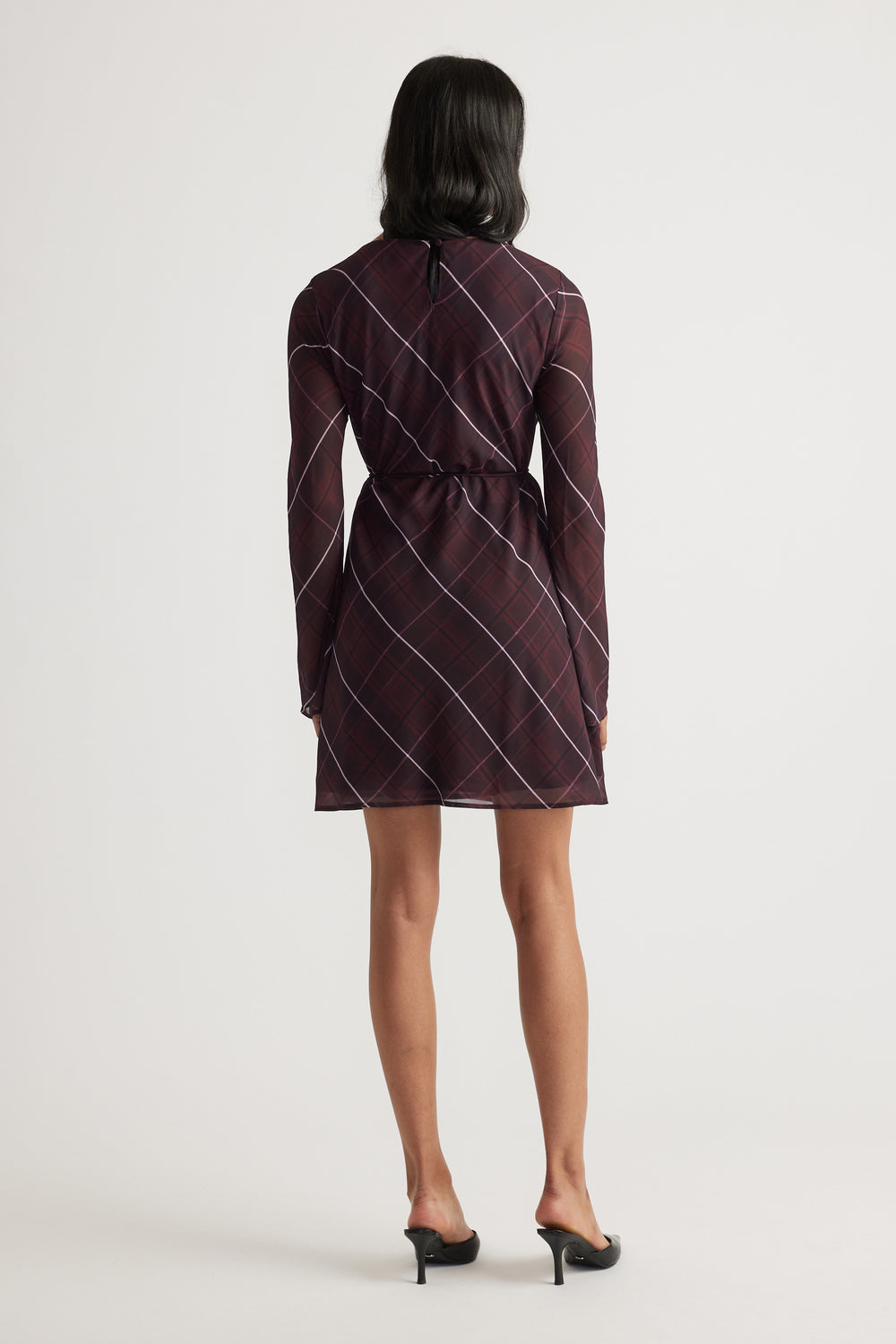 Woman wearing a long-sleeve dark checkered dress with a waist tie, shown from the back, paired with black heeled sandals