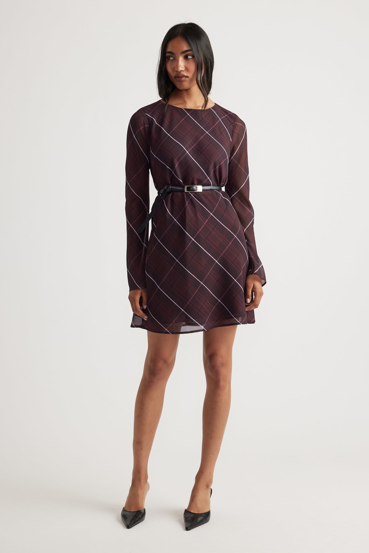 Woman wearing a long-sleeve plum dress with diagonal check print and a slim black belt, paired with black pointed heels