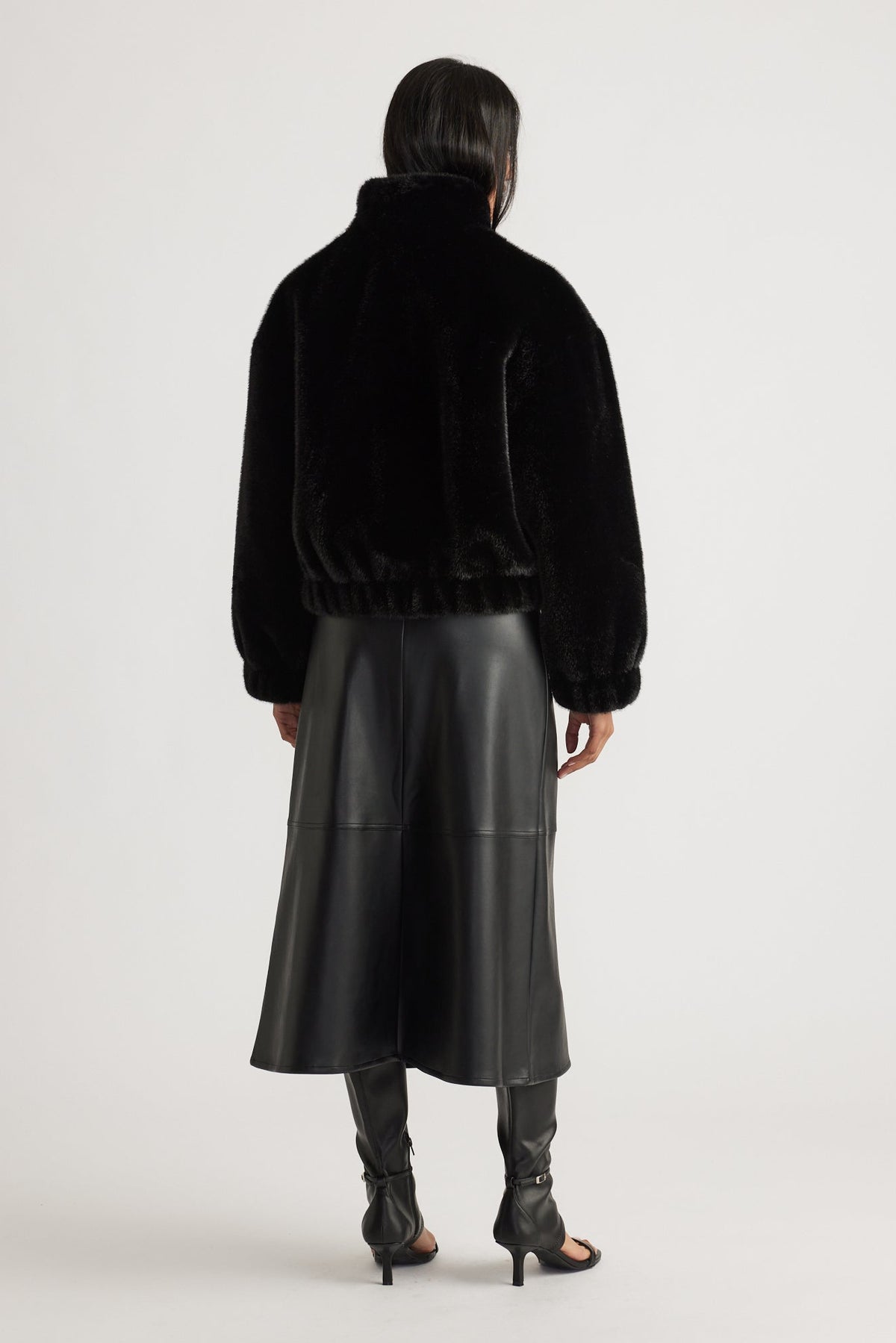 Woman wearing a black faux fur bomber jacket with elastic hem, paired with a sleek black leather midi skirt and heels.
