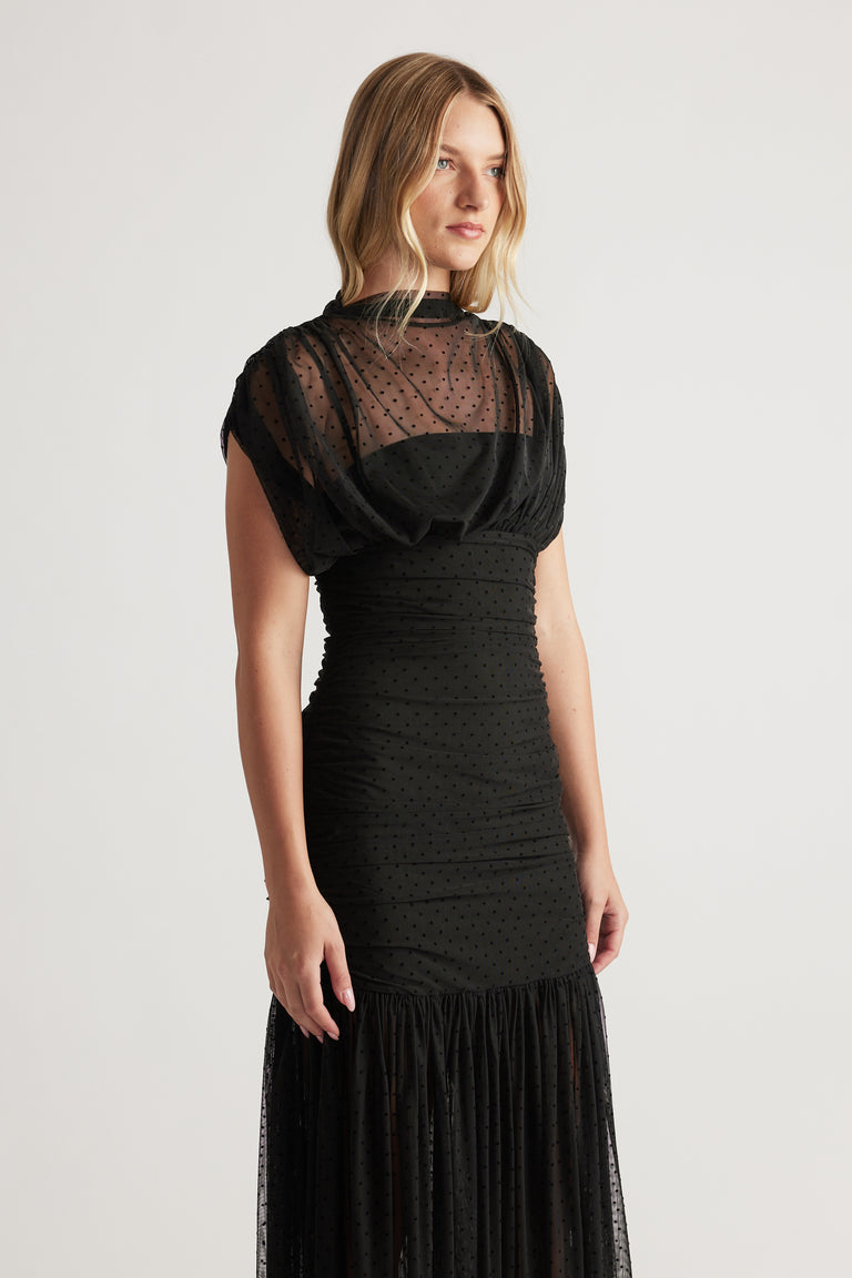 Woman wearing a fitted black mesh maxi dress with a high neck and subtle dotted pattern, featuring gathered details.