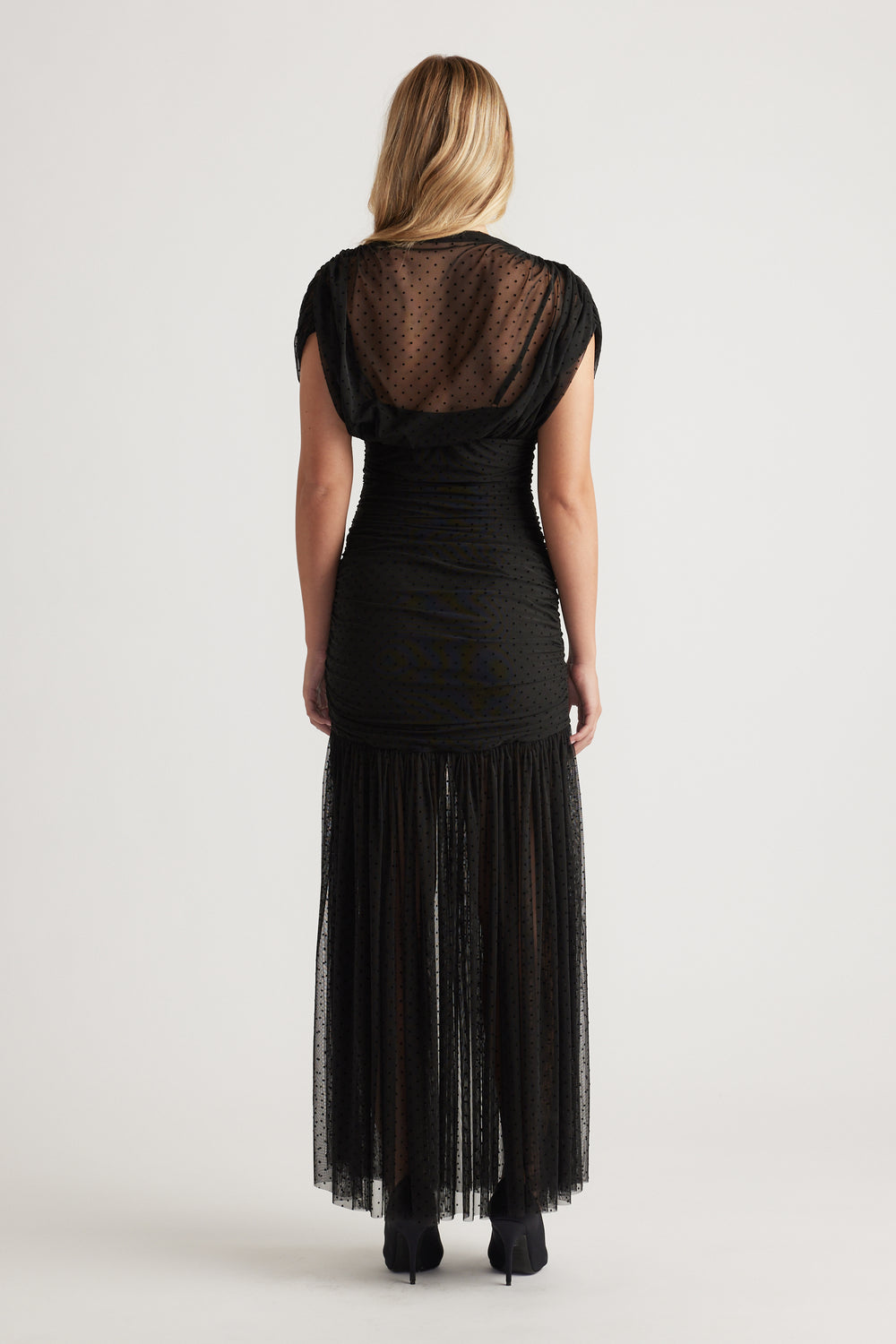 Woman wearing a black high-neck mesh maxi dress with subtle polka dots, ruched waist, and a tiered sheer skirt panel