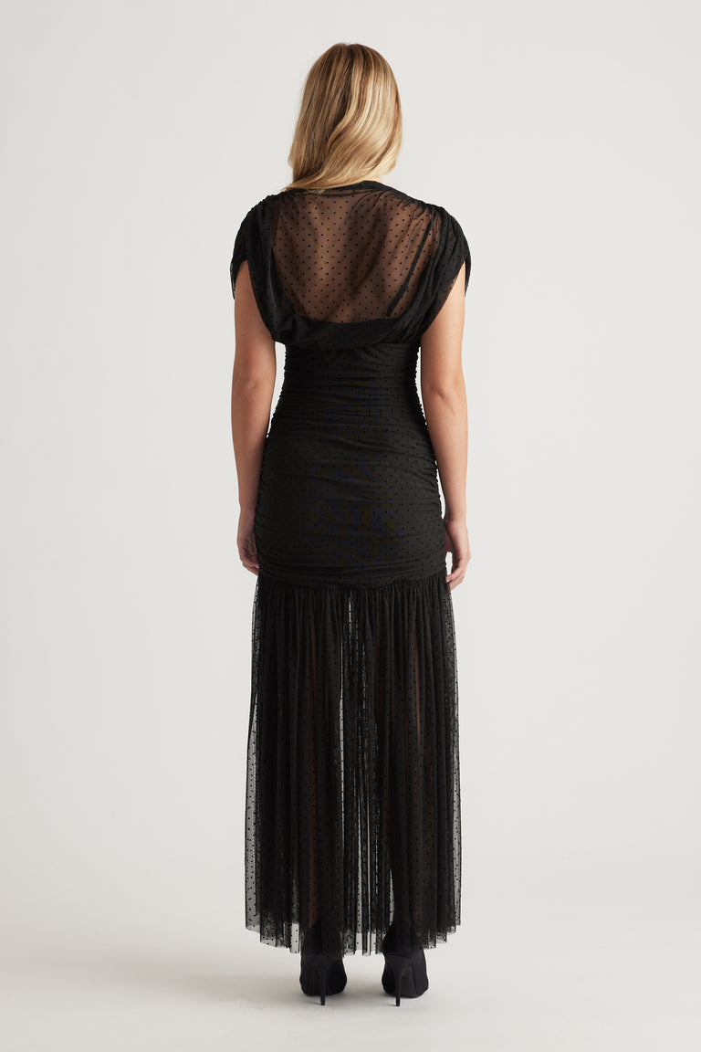 Woman wearing a black high-neck mesh maxi dress with subtle polka dots, ruched waist, and a tiered sheer skirt panel