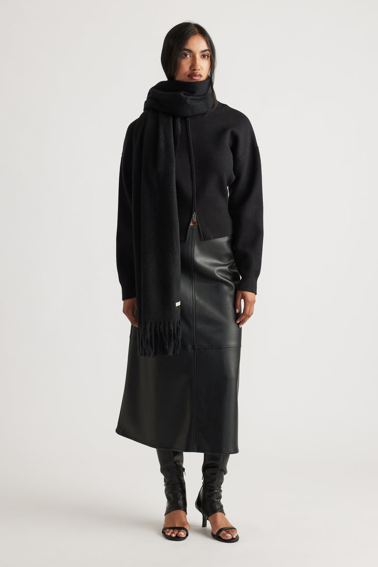Woman styled in a long black scarf wrapped around neck, paired with a fitted black sweater, faux leather midi skirt, and heels.