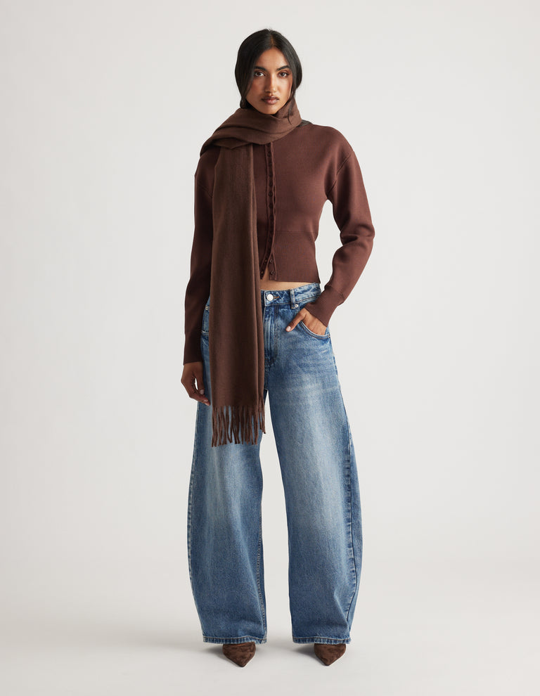 Woman wearing a long chocolate brown scarf draped over one shoulder with wide-leg faded blue jeans and pointed boots