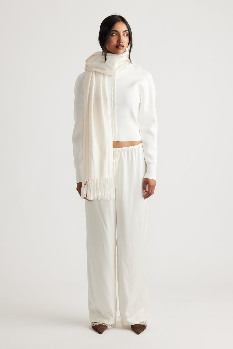 Woman dressed in an elegant all-white outfit featuring a soft, fringed cream scarf for a refined, cozy look