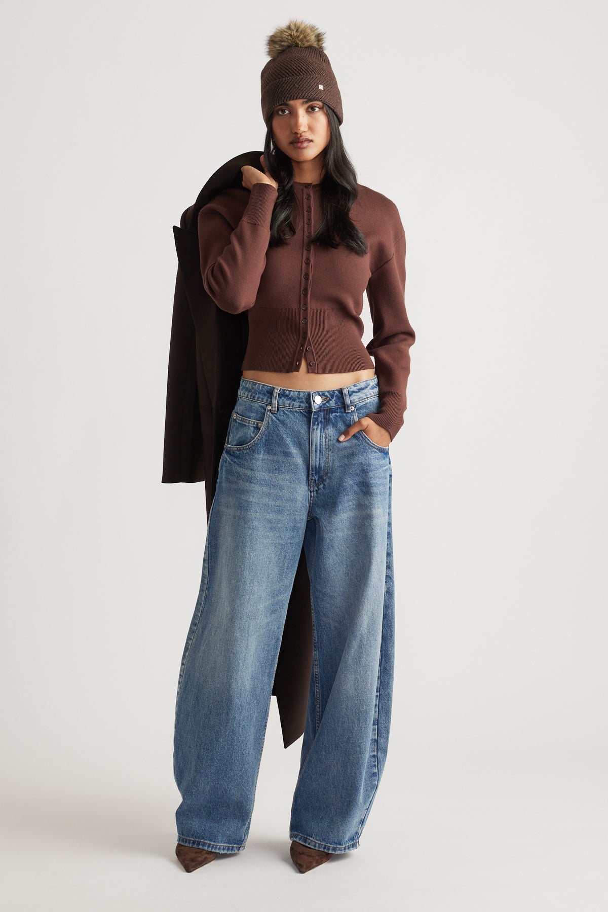 Woman wearing a fitted brown cardigan and wide-leg blue jeans, styled with a brown pom beanie and coat draped over shoulder