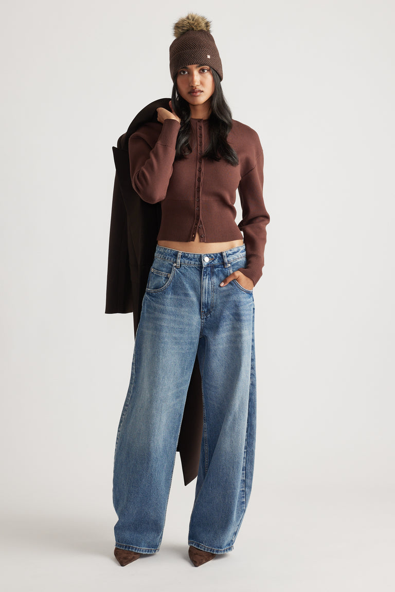 Woman wearing a fitted brown cardigan and wide-leg blue jeans, styled with a brown pom beanie and coat draped over shoulder