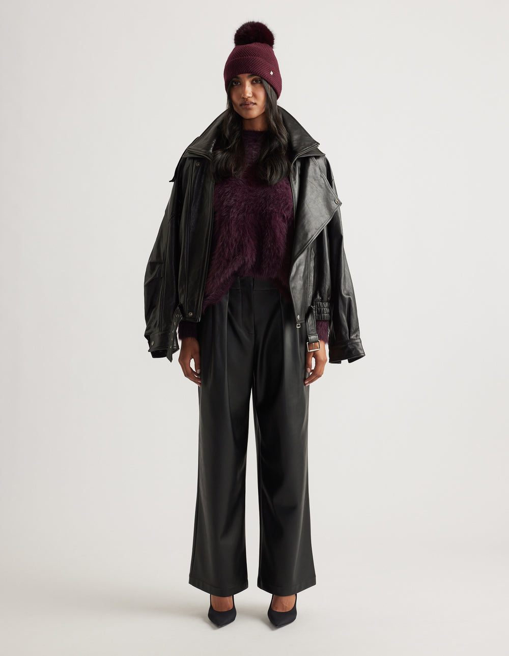 Woman wearing a plum knit sweater and matching pom beanie, styled with a black leather jacket draped over shoulders and wide-leg pants