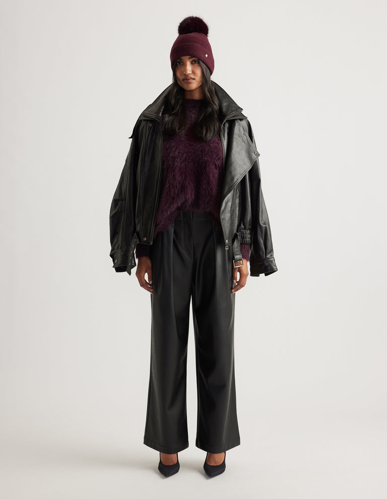 Woman wearing a plum knit sweater and matching pom beanie, styled with a black leather jacket draped over shoulders and wide-leg pants